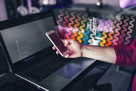 Alternative ADHD Treatment. Image of a laptop from an angled view with a person's hand hovering above it. The hand is holding a smartphone. Flowing from the phone, hand and forearm are squiggly lines in different colours.