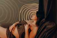 Postpartum depression. The photo shows a pregnant woman reclining, mostly covered by a dark, silky fabric. She appears to be resting peacefully, with her eyes closed, holding the fabric loosely over her body. The background consists of an abstract, wavy pattern of concentric lines, likely mimicking the look of topographic lines on a map. The colors in the image are soft and muted, with shades of dark brown, earthy green, and light beige from the pattern. The lighting is warm, enhancing the calm and relaxed mood of the scene.