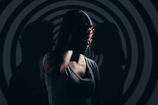 Psychedelic Therapy for Personality Disorders. The image features a woman with her hand covering part of her face, standing against a hypnotic, spiraling black-and-gray background. Her face is partially obscured, with light casting dramatic shadows that create a moody, intense atmosphere. Two silhouettes flank her, one on each side, as though they are shadows of her or representations of internal figures, adding an eerie, introspective feel. The woman's gray shirt and the subdued color palette, dominated by dark tones and soft contrasts, enhance the sense of depth and psychological tension. The concentric circles in the background draw the viewer's eye inward, amplifying a feeling of introspection and mystery.