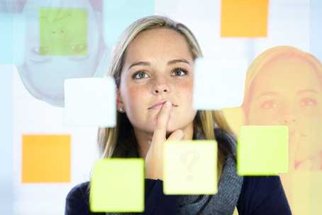 Are Psychedelics for Me? The image features a woman with blonde hair standing behind a transparent surface covered in colorful sticky notes, including shades of yellow, orange, green, and blue. She appears thoughtful, with her finger resting on her chin, suggesting she is reflecting or brainstorming. Her face is in focus, with a soft light illuminating her features. The composition includes blurred reflections of her face in the glass, adding depth and a dynamic visual element. The background is neutral, ensuring the sticky notes and her expression remain the focal points.