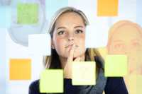 Are Psychedelics for Me? The image features a woman with blonde hair standing behind a transparent surface covered in colorful sticky notes, including shades of yellow, orange, green, and blue. She appears thoughtful, with her finger resting on her chin, suggesting she is reflecting or brainstorming. Her face is in focus, with a soft light illuminating her features. The composition includes blurred reflections of her face in the glass, adding depth and a dynamic visual element. The background is neutral, ensuring the sticky notes and her expression remain the focal points.