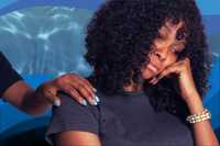 Search for Purpose. Image of a person from the shoulders up, sitting with their head resting on their hand. They are not smiling. They have brown skin, black curly shoulder-length hair, and are wearing a dark blue t-shirt. There is another person's comforting hand on their shoulder. The background is a blue water-like pattern.