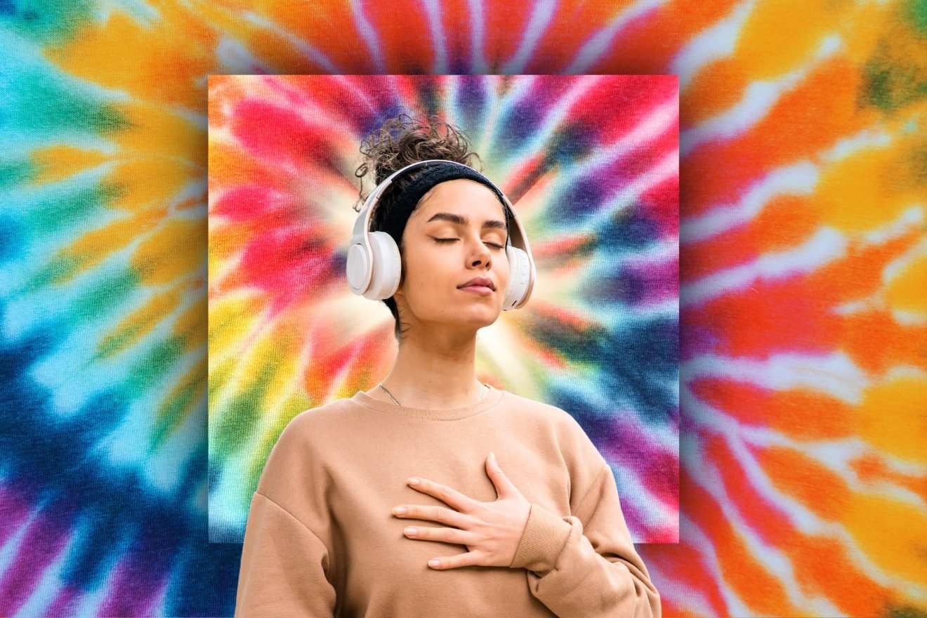 Psychedelic path. The image shows a woman with closed eyes, wearing white headphones, and placing her hand on her chest in a calm pose. She stands against a colorful, vibrant tie-dye background with swirling patterns of red, orange, yellow, and blue, giving the scene a peaceful yet dynamic feel. The composition emphasizes her serene expression against the energetic backdrop.