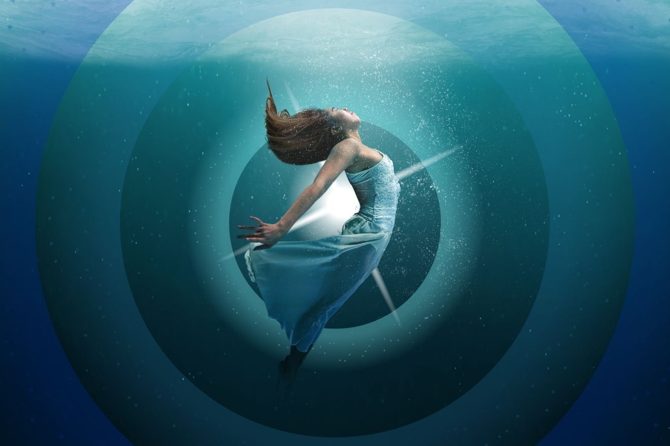 Healing with psychedelics. The image depicts a woman in a flowing white dress, suspended underwater. She is positioned in the center of the frame, with her body leaning backward and her hair flowing upwards. The background features a series of concentric circular patterns, giving a sense of depth and movement. The water is a gradient of deep blues and greens, with bubbles scattered throughout, contributing to the fluid, dreamlike atmosphere. The overall composition highlights the subject’s graceful motion within the aquatic environment.