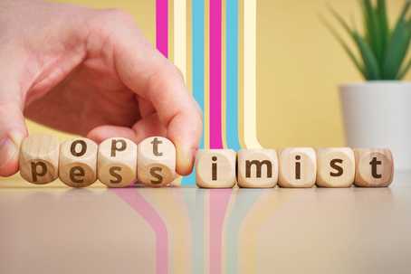 Pessimism Bias. A row of dice with letters on them. The letters spell out "pessimism," but someone's hand is busy rotating the dice to show that it spells out optimism instead. There is a potted plant in the back right corner and some stripes along the light yellow wall in hot pink, cream, and turquoise.