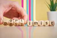 Pessimism Bias. A row of dice with letters on them. The letters spell out "pessimism," but someone's hand is busy rotating the dice to show that it spells out optimism instead. There is a potted plant in the back right corner and some stripes along the light yellow wall in hot pink, cream, and turquoise.